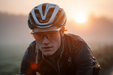 Focused cyclist in rain-splashed jacket and helmet riding into sunrise, determined close-up portrait capturing grit and endurance