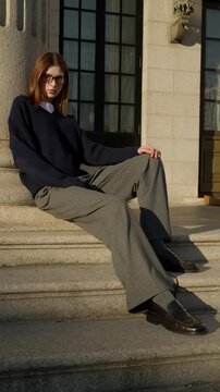 Stylish fashion model wearing classic outfit posing on stone steps near historic building during warm sunset light