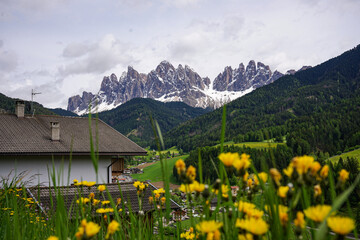 Alpine village with traditional houses, green hills and dramatic rocky mountain peaks in the background. Spring countryside landscape and nature travel concept.