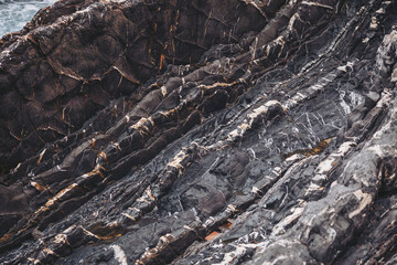 The scene shows various layers of dark rocks on the shore with cracks and lines. Waves crash gently against the edges near the ocean. Sunlight highlights many details on the rock surface