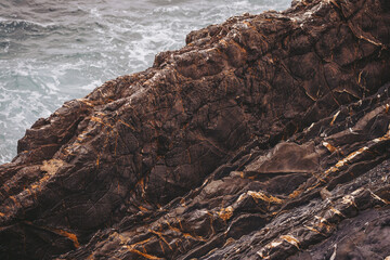 Rocks are seen against the backdrop of ocean waves crashing against the shoreline. The scene captures the texture of the rocks and the movement of the water during the afternoon