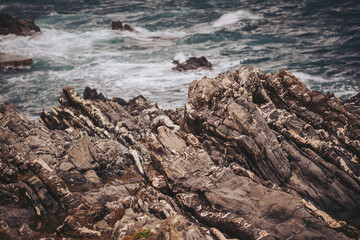 Waves crash against the rocky shoreline while clouds fill the sky above. The scene shows rough rocks and foamy water, capturing the coastal landscape during overcast weather