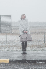 Young woman in a gray coat holding a large stylish bag standing on a road during a snowfall. Winter fashion and lonely travel concept.