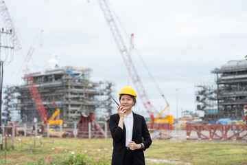 An Asian female engineer wearing a safety helmet is using a radio communicator at an industrial plant construction site, surrounded by heavy machinery and cranes, managing modern industrial operations