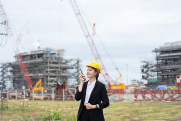 An Asian female engineer specializing in power plant construction, communicating with her team via radio, managing safety, coordinating machinery, and overseeing ongoing industrial development.