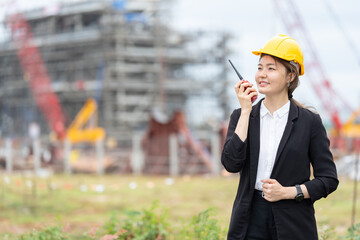 A female engineer in work uniform and safety helmet in a large industrial plant, using communication devices to control machinery, symbolizing innovation, industrial growth, and management efficiency.