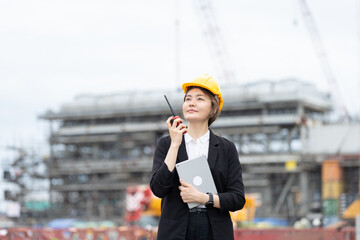 An Asian female engineer, wearing a helmet and carrying a radio, works on a construction site, overseeing heavy machinery and structures, leading and implementing modern industrial practices.