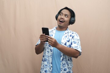 Excited Southeast Asian man wearing headphones, blue t-shirt, and patterned shirt while holding smartphone, enjoying music or audio content for lifestyle and advertising concepts.