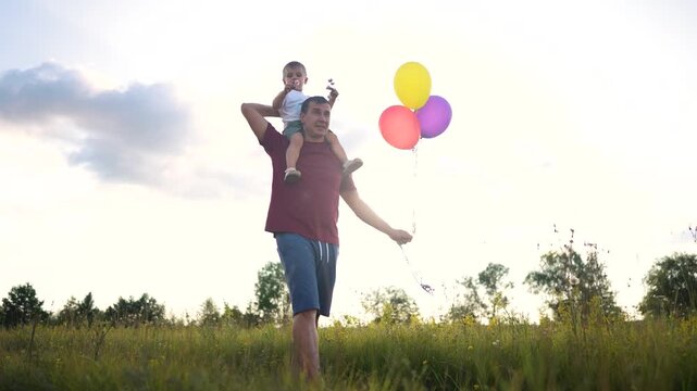 Father and son in field holding balloons. Balloons with a man and boy. A woman is walking outdoors with her shoulder bounding in the field. A dad and his son are in a lifestyle field carrying.