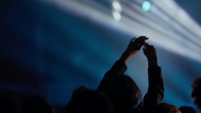 Dark crowd silhouettes raise hands and phones celebrating peak live music moment under strobe lighting and intense concert atmosphere