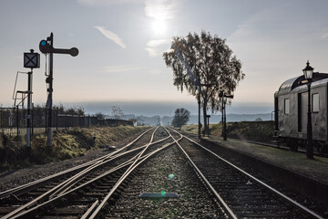 Railway tracks diverging at a switch with old signals and a vintage carriage, silhouetted against a bright backlit sun and misty autumn sky.