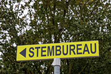 Bright yellow sign with the text Stembureau and an arrow, indicating a polling station for elections in the Netherlands, against green trees.