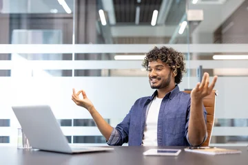 Fotobehang Lotusbloem A young Indian man sits at a desk in the office in a lotus position, resting and meditating  © Tetiana