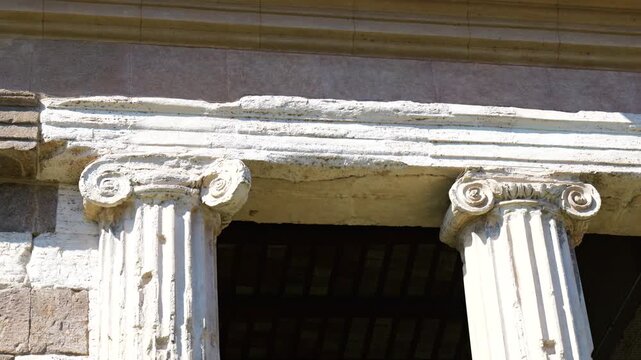two weathered ionic columns worn volute capitals support cracked entablature contrasting patched stone walls shadowed interiors ancient structure rome italy roman architectural detail 