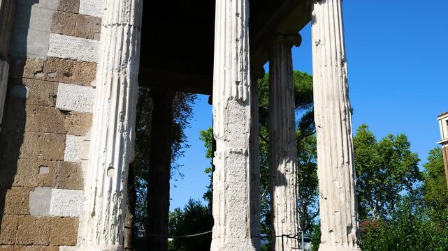 weathered ionic columns patched stone walls temple portunus stand beneath clear blue sky glimpses greenery visible through colonnade rome italy of 