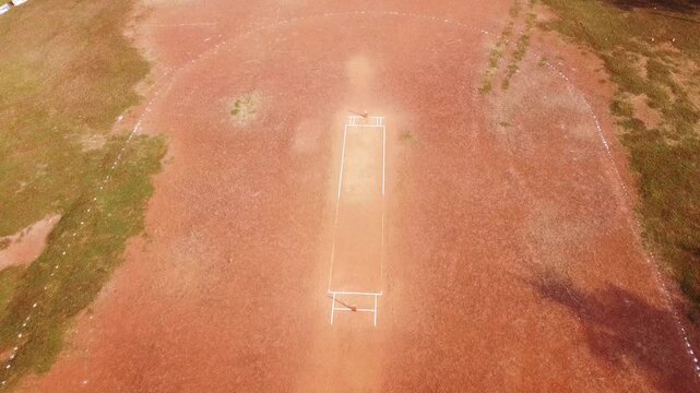 Top-down aerial drone shot of a rural cricket pitch prepared on red clay soil, showing white crease markings and metal wickets in a dusty, open field setting.
