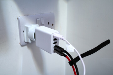 A white electrical adapter is plugged into a wall socket, connected through a power converter. Three USB ports on the adapter are in use with attached cables. The wall plate shows a UK-style socket.