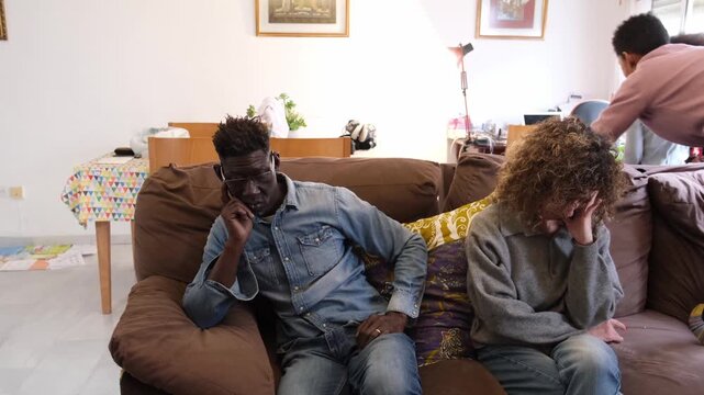 Stressed interracial parents sitting on the sofa feeling tired and overwhelmed while their energetic son and daughter run and play noisily around them