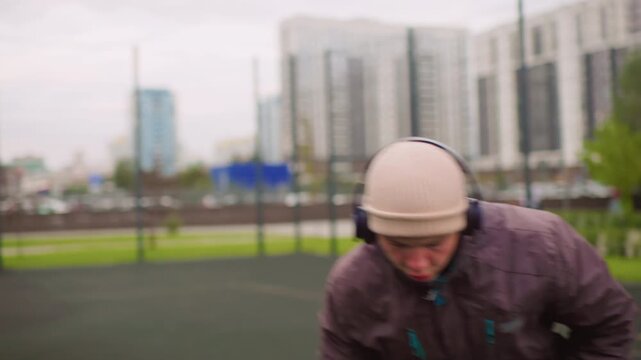 Caucasian man dribbling basketball on court, wearing beanie and headphones, padded jacket and sneakers, performing low crossover and focused footwork on rubber surface, city highrises in background,