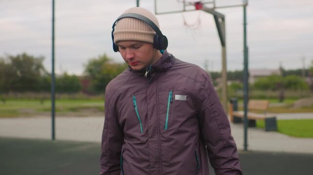 young man on basketball court wearing headphones, layered jacket and beanie, casually dribbling basketball, posing for portrait under overcast sky, hoop and park in background, relaxed urban sports