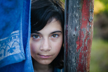 Close-up portrait of a young girl looking through a narrow gap between a blue fabric and a weathered wooden post, with soft natural light and an intense, thoughtful gaze.