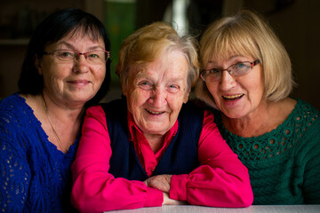 Warm portrait of three elderly women sitting close together, smiling at the camera, showing friendship, family bonds, and shared joy in a cozy indoor setting.