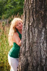 Blonde woman peeking from behind a tree in a green summer meadow, captured in soft natural light, with a playful yet calm expression and a relaxed outdoor atmosphere.