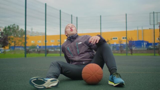 young white man seated on basketball court wearing beanie and jacket, holding ball while resting after practice, fenced city park backdrop with yellow warehouse and overcast sky, sneakers and turf
