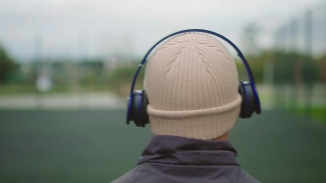 person with beanie headphones facing field, back turned to camera in urban park, blurred sports ground beyond, cool overcast light, wool jacket and wireless headphones signaling music listening, mood