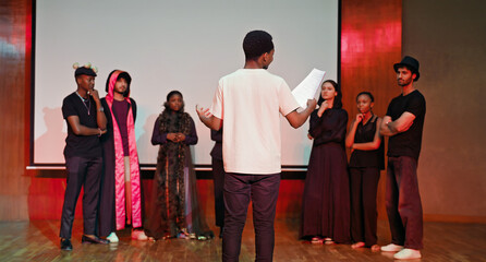 African young adult gen z male man hold scripts paper read play story lines scene standing indoor stage hall. Happy group boy girl clap hand enjoy do work show drama practice day inside campus place