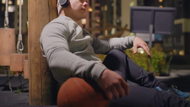 nighttime urban bench man with basketball, wearing headphones and beanie, leaning against wooden post under lamplight, city skyline bokeh, quiet contemplative mood, casual sweatshirt and sneakers,