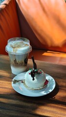 Coffee beverage in a takeaway plastic cup served alongside a small dessert on a wooden table, illuminated by warm natural sunlight. The cozy cafe atmosphere and soft shadows create a relaxed lifestyle