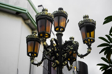 Ornate Vintage Street Lamp with Multiple Lanterns and Decorative Metal Details, Low Angle View Against Urban Building and Overcast Sky
