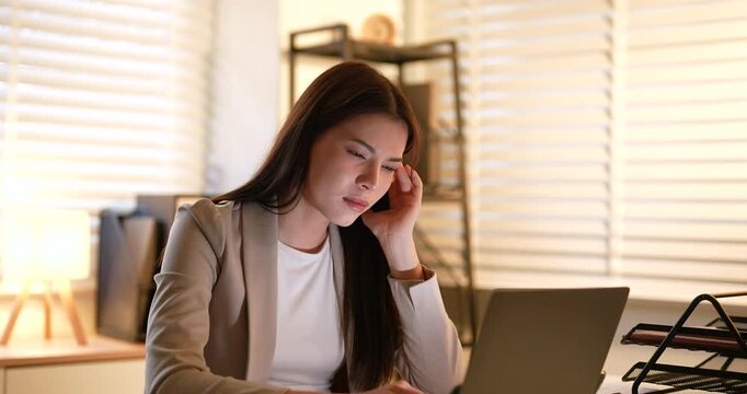 Asian businesswoman holding head while working on laptop at night, overwhelmed with workload and deadline pressure, concept of tired employee stress management and overwork in corporate environment