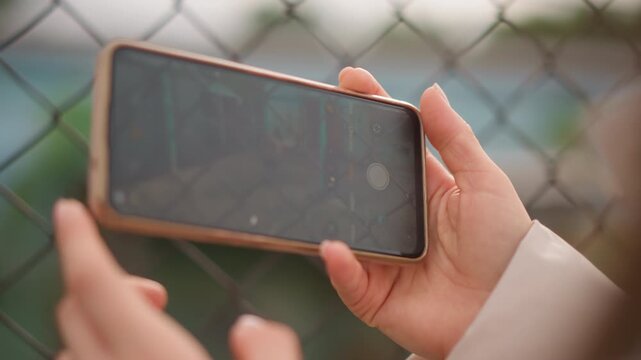 hands holding phone with map, closeup of person checking route against chainlink fence, touchscreen gestures zooming into satellite map, urban background blurred, soft warm light, focused on device