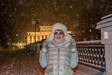 Mature adult female in winter clothes on the background of old town with night illumination during of snow fall.