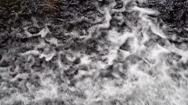 Overhead close up view of turbulent river water surface flowing rapidly over a dam. Rushing stream creating white foam, bubbles, and abstract textures. Concept of hydro energy, power, and nature backg