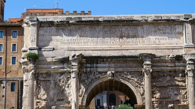 frontal daytime richly carved ancient roman triumphal arch latin inscriptions relief sculptures frame archway amid historic buildings rome front view italy empire emperor 