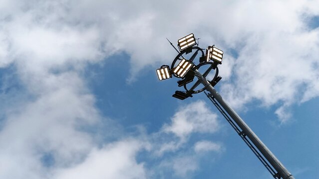 High mast lighting pole with multiple LED floodlights against a bright cloudy sky background.