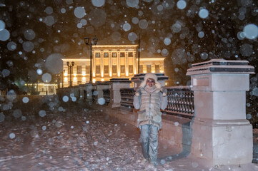 Mature adult female in winter clothes on the background of old town with night illumination during of snow fall.