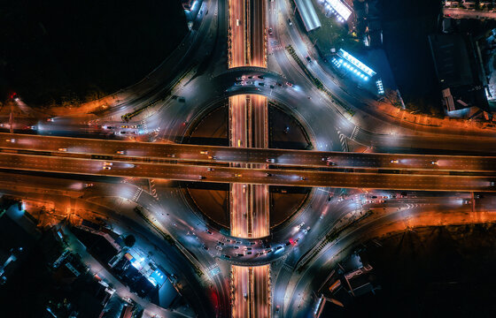 Stunning top-down aerial view of a complex highway interchange at night, featuring vibrant long-exposure traffic light trails in Nonthaburi, Thailand.