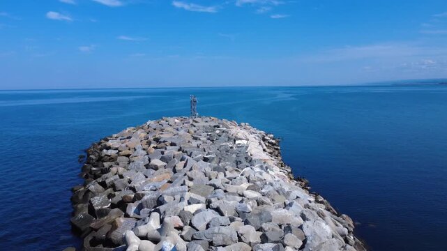 Flyover of a rock breakwater to its end, topped by a metal lighthouse pointing towards a calm ocean merging with the blue sky on the horizon. St. Lawrence River, Matane, Quebec, Canada, 2025.