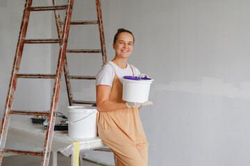 A woman smiles while holding a paint bucket next to a ladder in a freshly prepared room. She is wearing an apron and preparing to start painting the walls