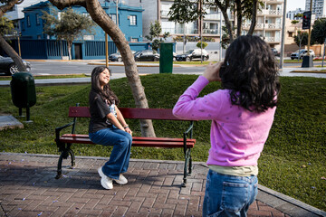 A teen is taking a picture of a girl sitting on a bench