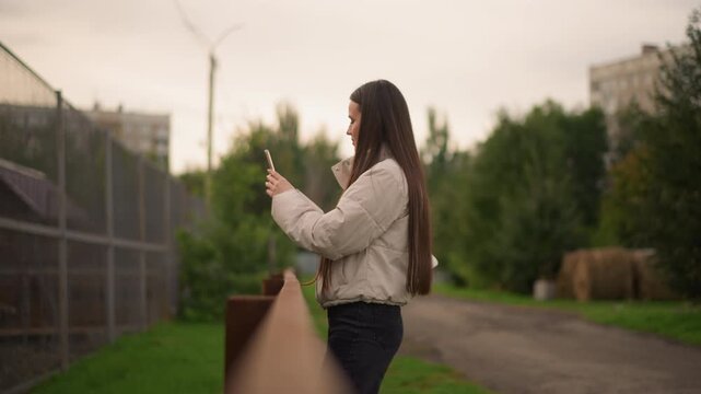 Young woman texting outdoors near fence, suburban path with apartment blocks and trees, overcast sky, quilted jacket and braided hair, casual student checking messages while leaning on railing, soft