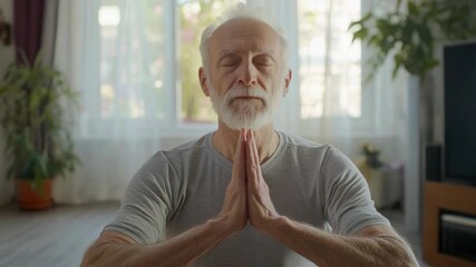 A man seated cross-legged on the floor, eyes closed, in a relaxed meditative state. His hands are clasped together in front of him. The room is well lit, suggesting it's daytime.