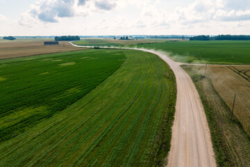 Obraz premium Aerial view shows pale gravel road curving through crop rows, faint dust plume, utility poles, lone barn, and trees under broken clouds in daylight across farmland.