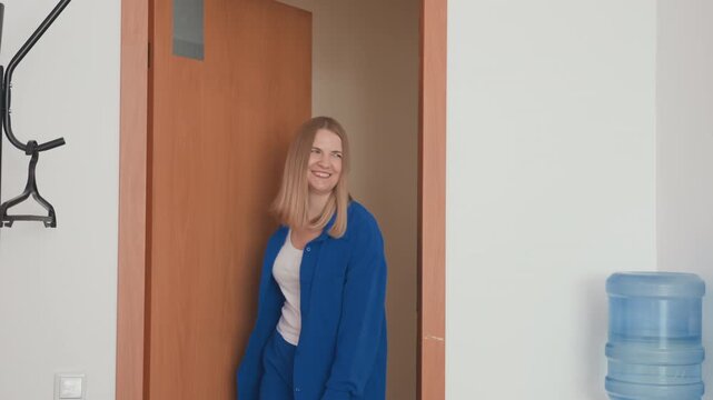 Woman enters office doorway beside cooler, blonde woman in blue blazer peeks and smiles at colleague reception area with coat rack and bright neutral walls casual greeting, candid workplace