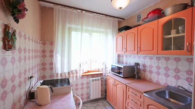 cozy kitchen with orange cabinets, patterned tile walls, a stainless steel sink, microwave, electric stove, sheer curtains, and a radiator under the window. Compact and functional