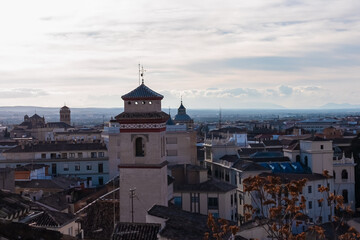 Obraz premium High angle view highlights a historic Mudejar church tower rising above traditional tiled rooftops against a backdrop of distant mountains and a cloudy sky in the Albaicin district of Granada, Spain.